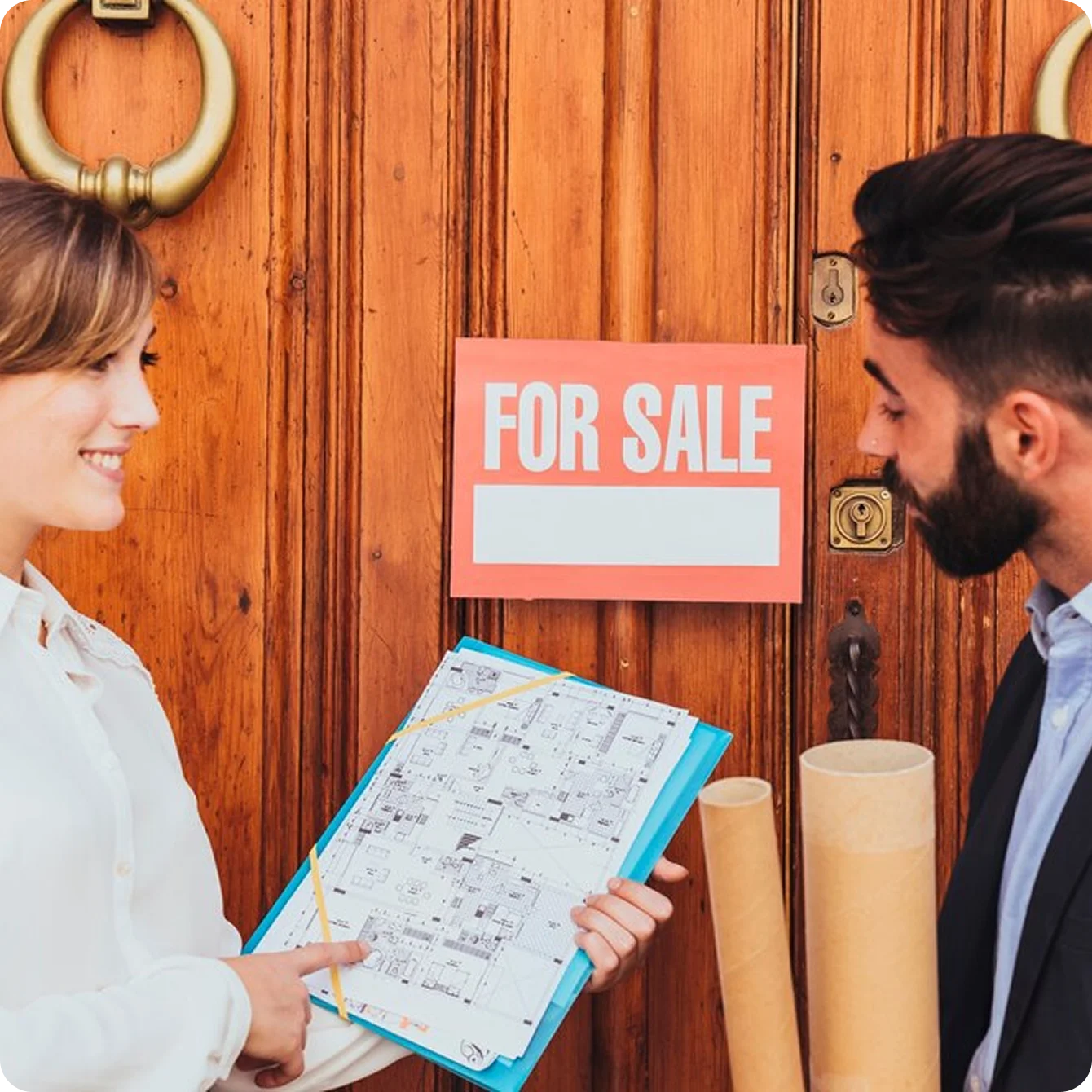 Smiling woman showing architectural floor plans to a man standing near a wooden door with a 'For Sale' sign.
