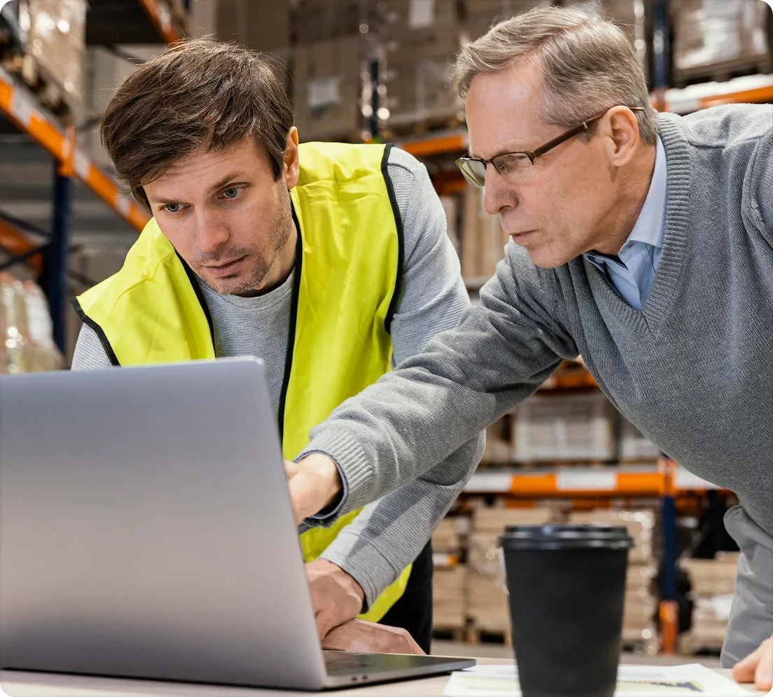 Two men checking a laptop in a warehouse, one wearing a high-visibility vest
