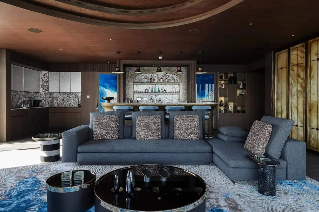 Modern living room with a large sectional sofa, circular coffee tables, a rug with blue accents, and a marble bar area in the background.