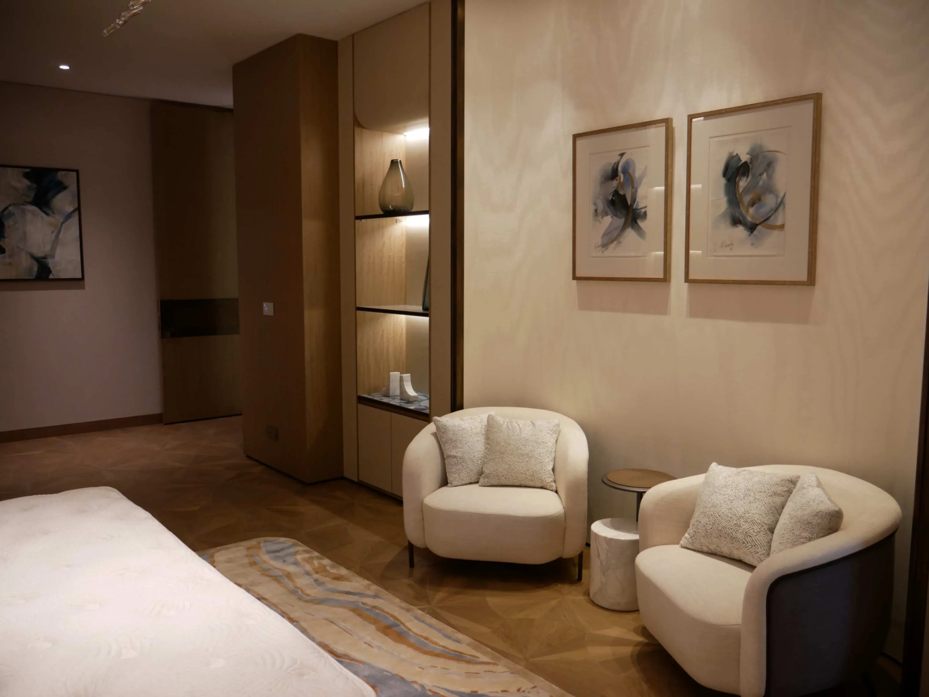 Modern bedroom corner with two cream armchairs, a small side table, abstract wall art, and built-in shelving with decorative vases.