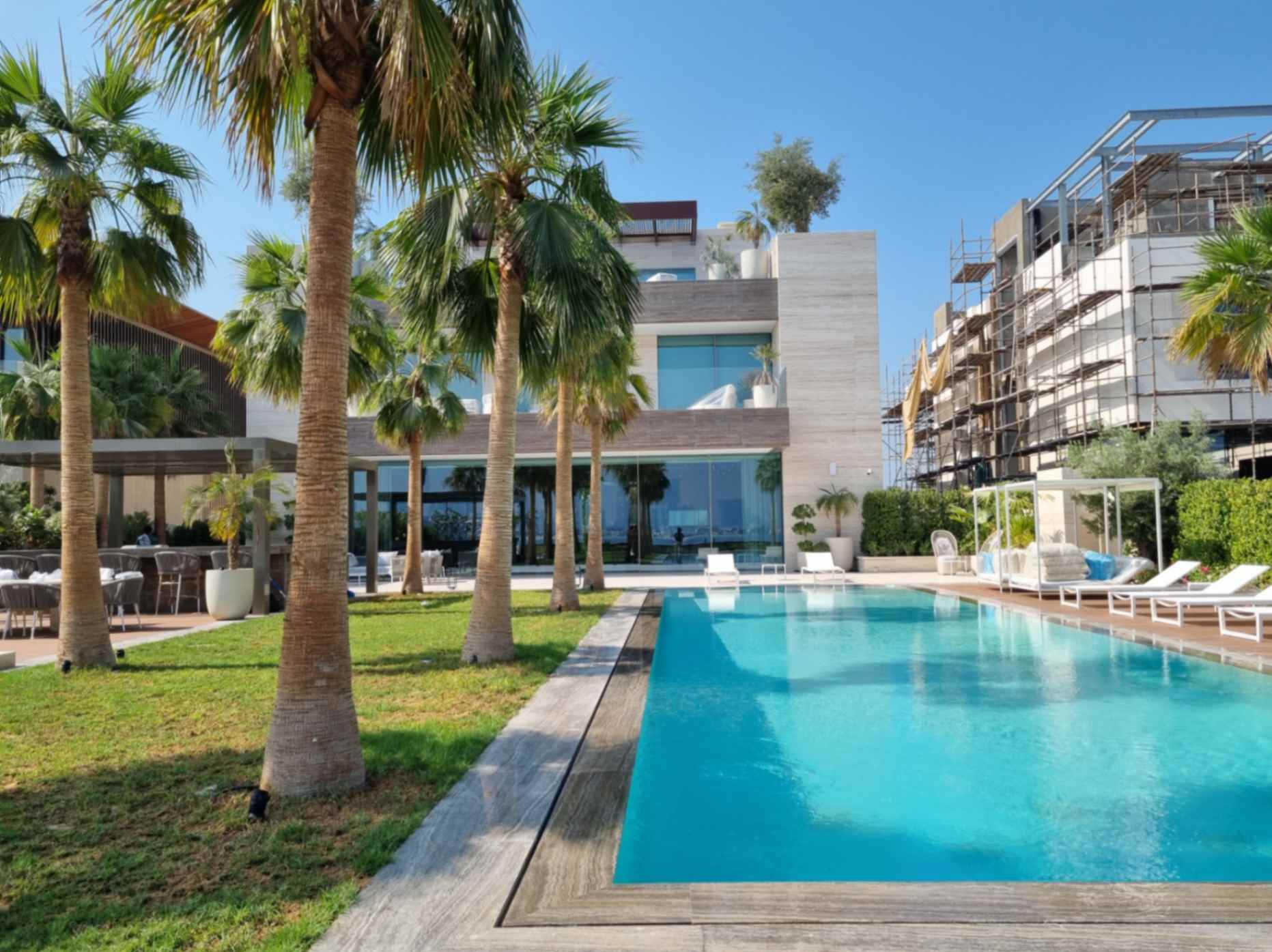 A sleek modern villa featuring a swimming pool surrounded by lush palm trees under a clear blue sky.