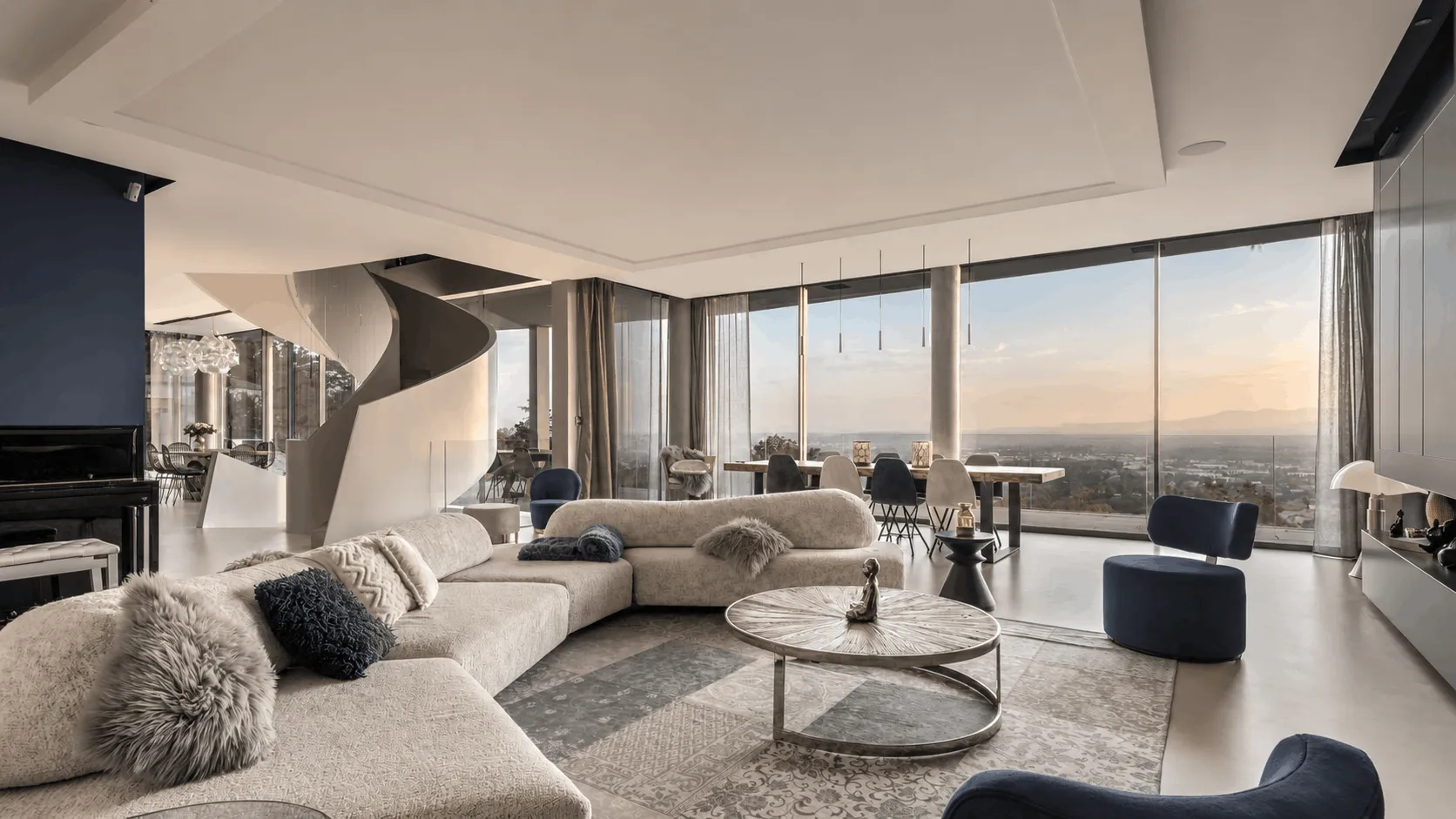 Salon lumineux avec canapé bleu, table basse en verre, tapis coloré, mur bleu foncé, salle à manger en bois clair, et grande fenêtre donnant sur un arbre.