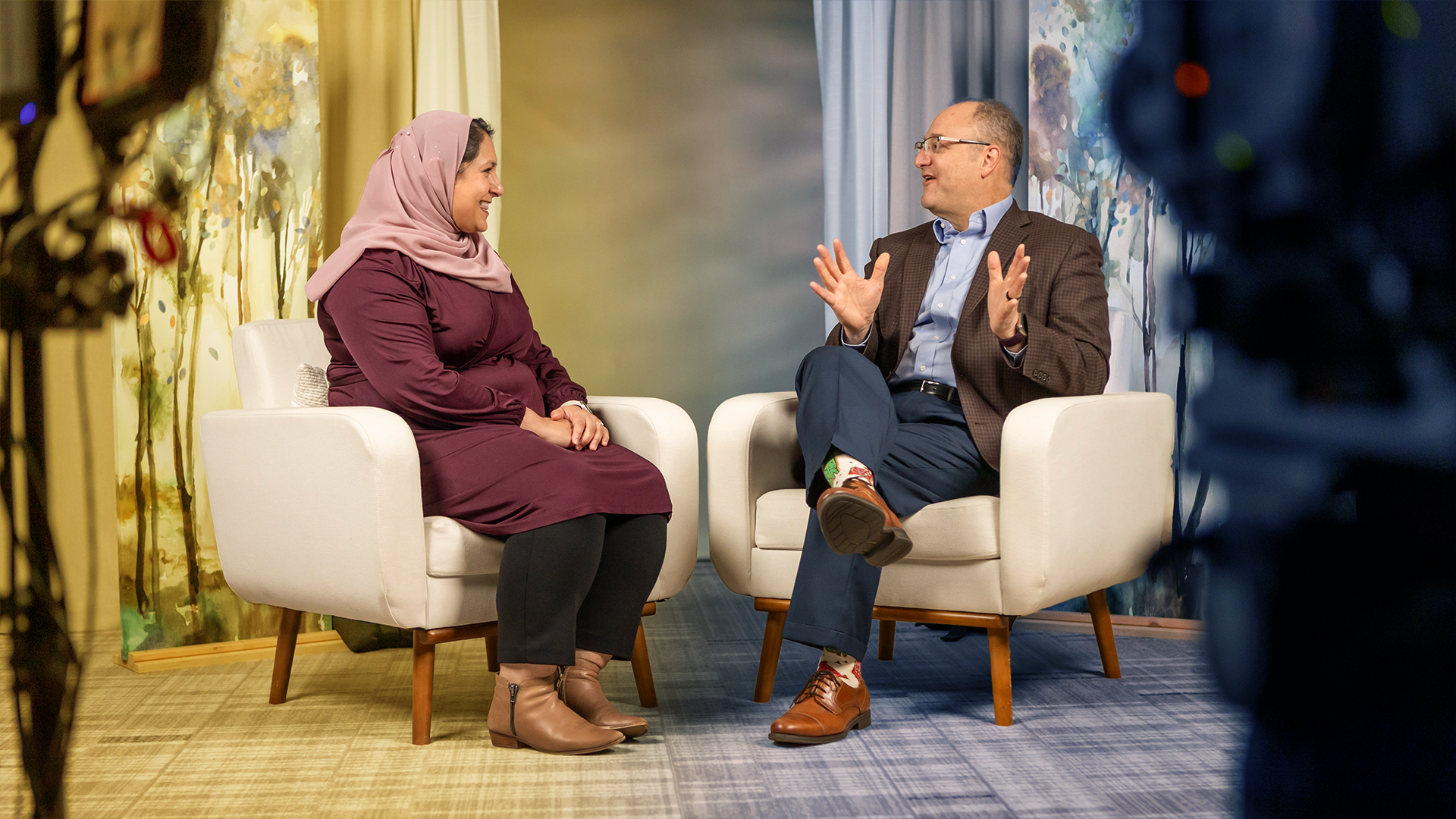 Two people sitting in white armchairs having a conversation in a warmly lit room with artistic wall decor.