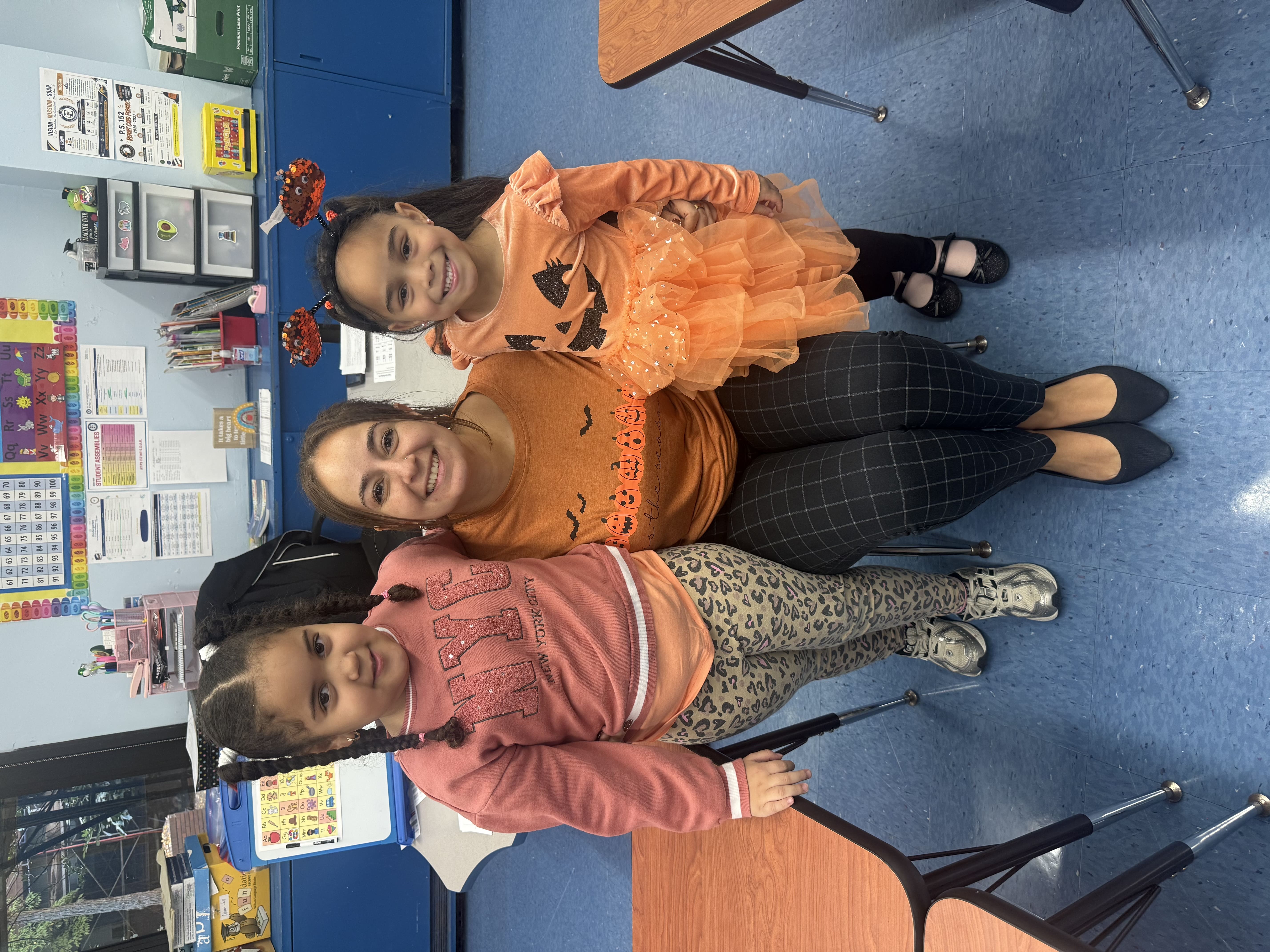 Two PS 152 students in orange pumpkin outfits with their teacher for Pumpkin Day