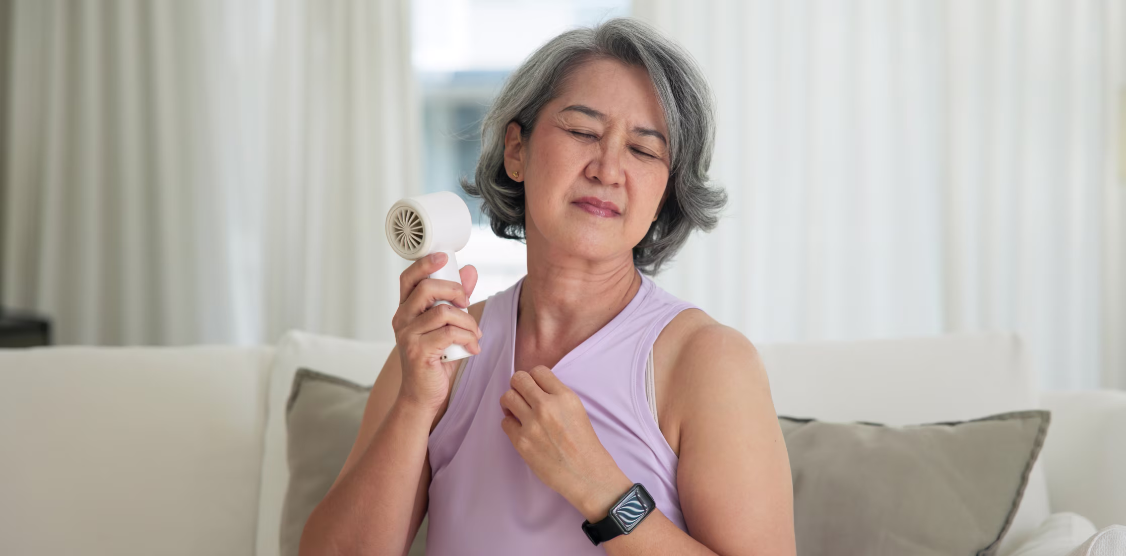 Menopausal woman on sofa using a handheld fan to relieve a hot flash, a symptom reported by 80% of women.