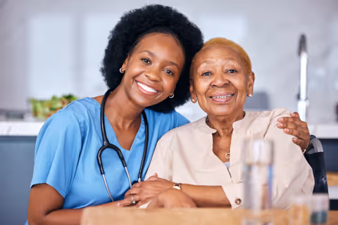 Smiling female healthcare worker in blue scrubs with stethoscope hugging an elderly woman in a wheelchair.