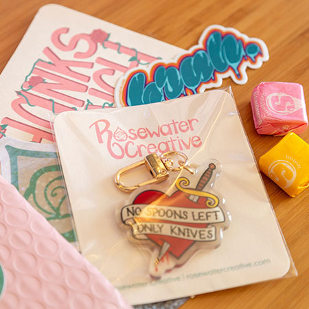 A photo of a pink bubble mailer with a green circular sticker on it, open and appearing to spill out its contents. These include an acrylic keychain on a backing card in a cellophane sleeve, a die-cut sticker, a Rosewater Creative logo sticker, a thank-you card, and two Starburst candies laid out on a bamboo tabletop.