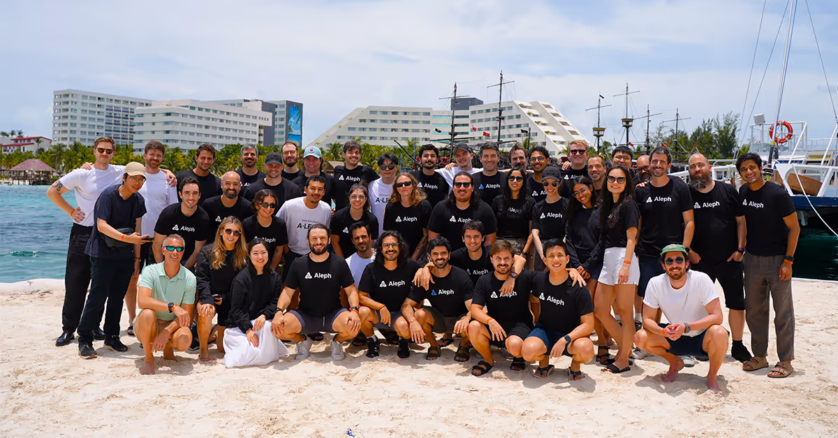 Large group of employees wearing Aleph t-shirts posing on a sandy beach in Cancun with hotels, ships, and blue sky in the background.