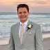 Smiling man in a gray suit with a white boutonniere standing by the ocean at sunset.