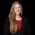 Smiling woman with long light brown hair wearing a black blazer and red top against a black background.