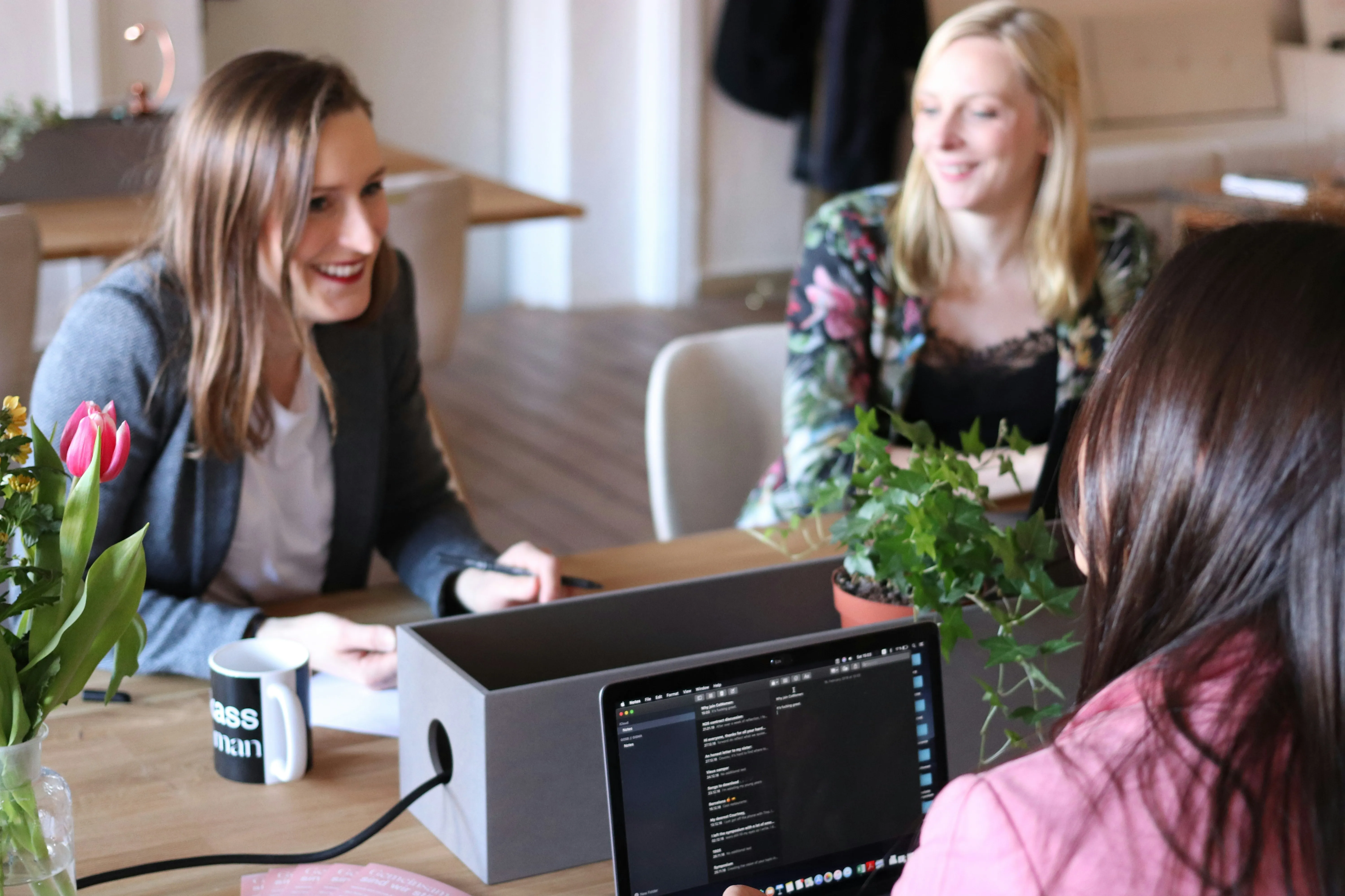 Three women engaged in a meeting around a table with a laptop, coffee mug, tulip flowers, and a potted plant.