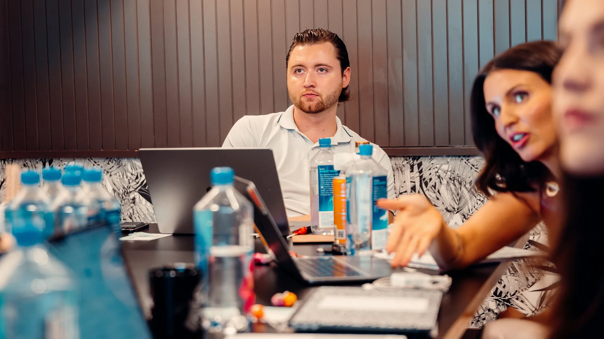 Man in a white polo shirt sitting at a table with laptops and water bottles, listening attentively while a woman gestures and speaks.
