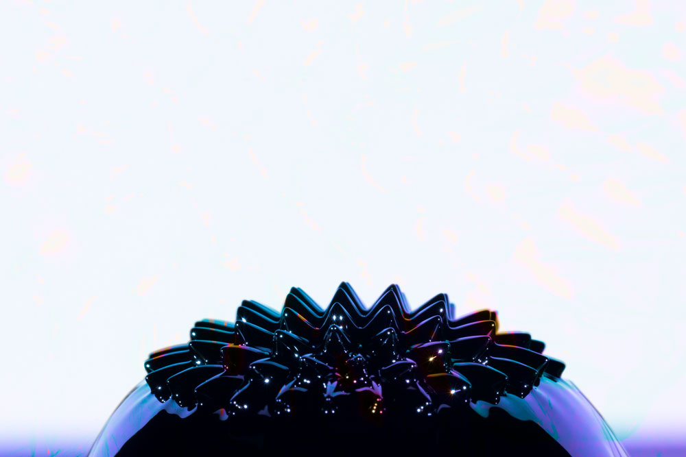 Close-up of a black ferromagnetic liquid metal forming sharp, star-like spikes on a smooth surface against a light background.