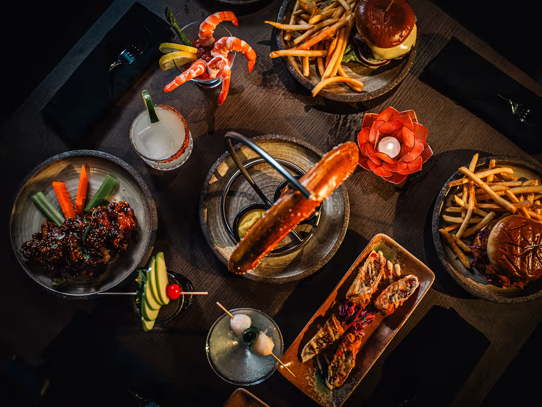 Overhead view of food and drinks at Dream 360, featuring burgers, fries, shrimp cocktail, wings, sushi rolls, and colorful cocktails on a table.