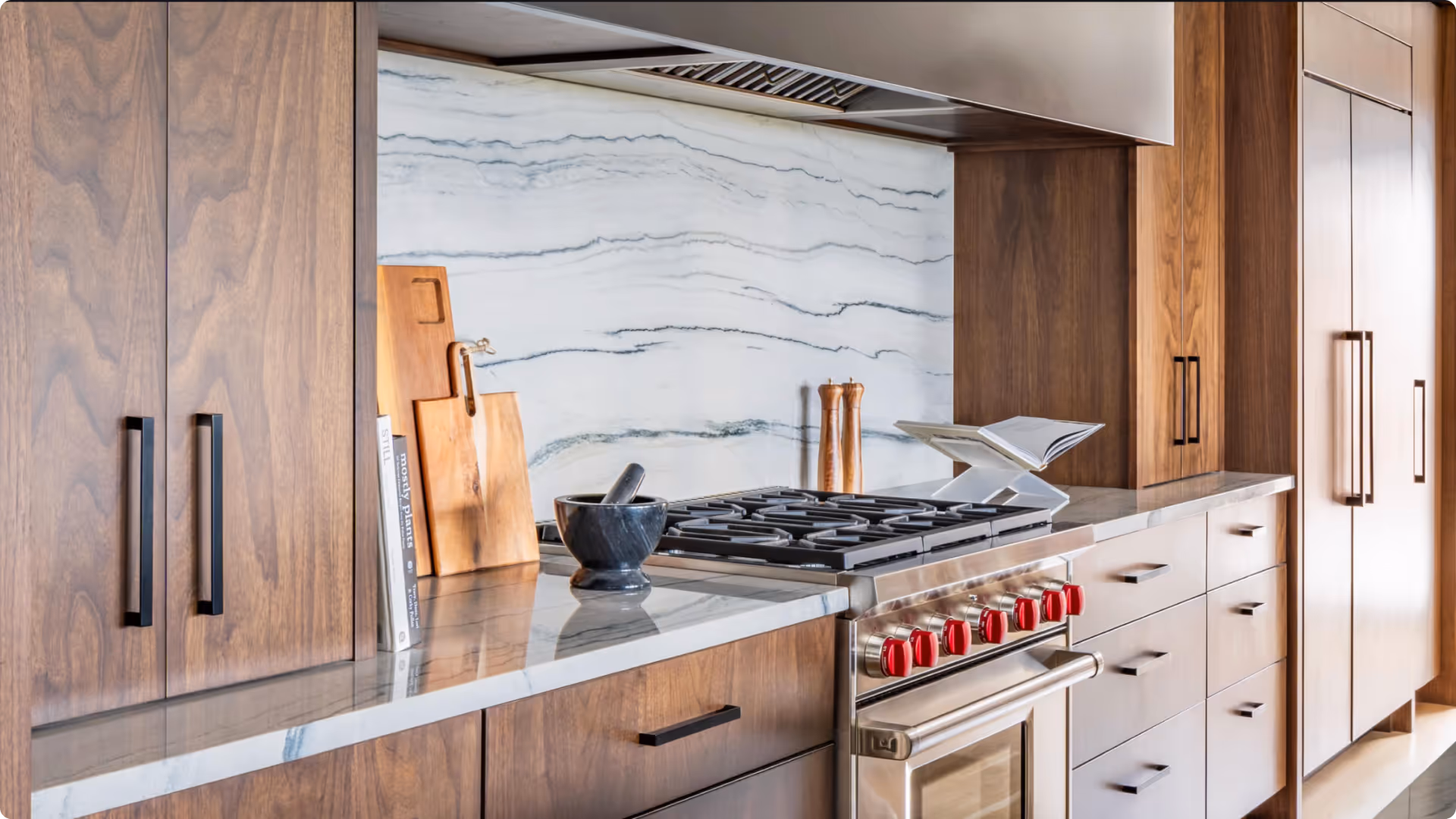 Modern kitchen countertop with wooden cabinets, a stainless steel stove with red knobs, marble backsplash, cutting boards, mortar and pestle, salt and pepper shakers, and a cookbook stand.