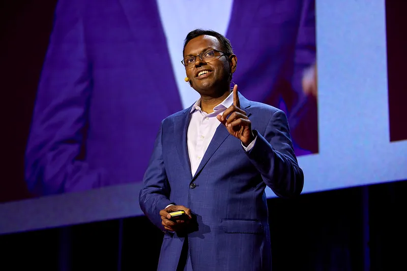 Man in a blue suit speaking on stage with a microphone headset and raising one finger.