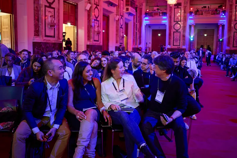 A diverse group of people seated in an ornate conference hall with red carpet, engaged in conversation and smiling.