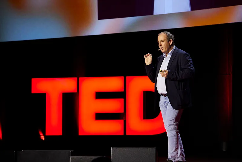 Man in blazer and light pants speaking on stage in front of large red TED letters.