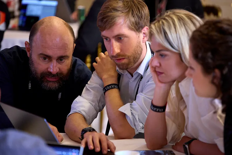 Four people closely collaborating and looking intently at a laptop screen in a conference setting.