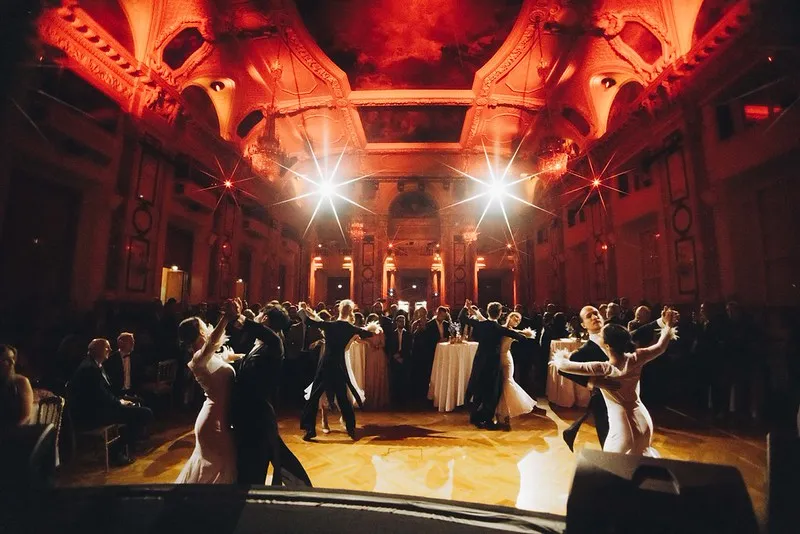 Several couples dancing elegant ballroom dances in a grand, ornately decorated hall with bright overhead lights.