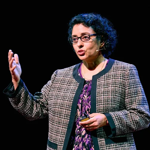 Woman with curly hair and glasses speaking and gesturing on stage wearing a patterned jacket and microphone headset.