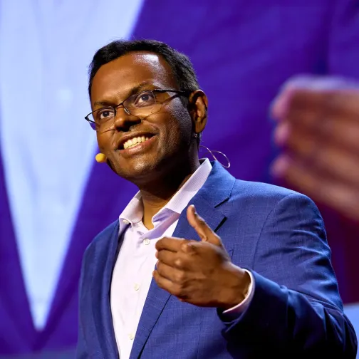 Man in blue suit and glasses speaking on stage with a headset microphone and gesturing with his hand.