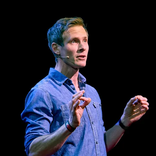 Man in a blue denim shirt speaking with hands raised and wearing a headset microphone.