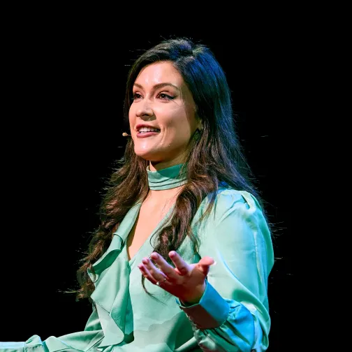 Woman with long dark hair wearing a light green blouse speaking and gesturing on stage with a black background.