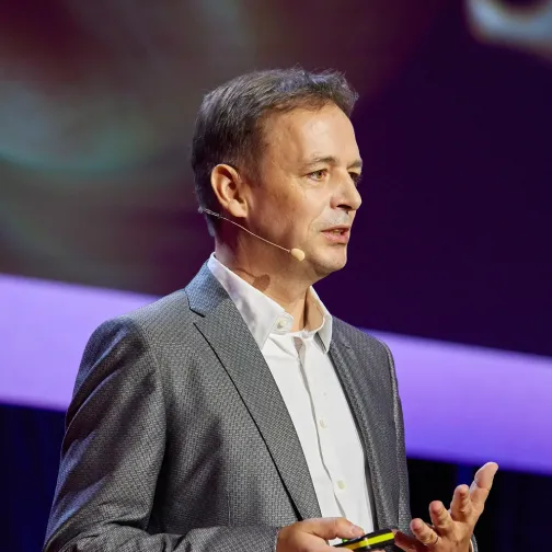 Man wearing a gray suit and white shirt speaking on stage with a headset microphone and gesturing with his hand.