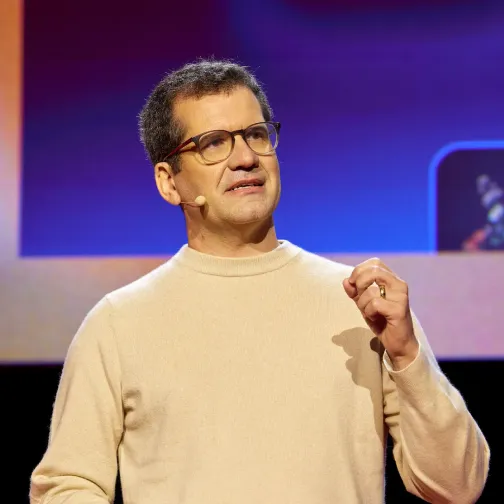 Man with glasses and beige sweater speaking on stage with a microphone headset.