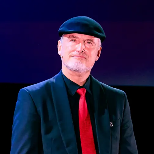 Man wearing a black beret, black suit, black shirt, and red tie against a dark background.