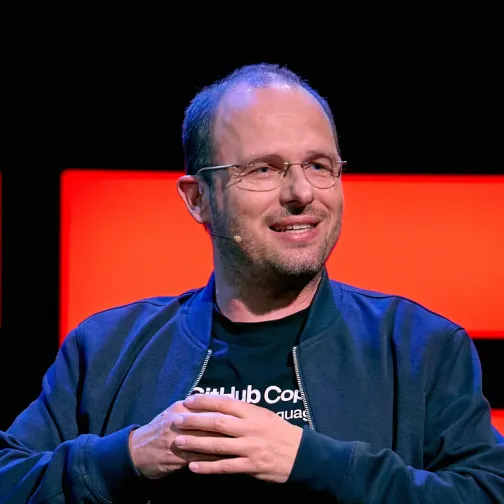 Man wearing glasses and a dark jacket speaking on stage with a red background.