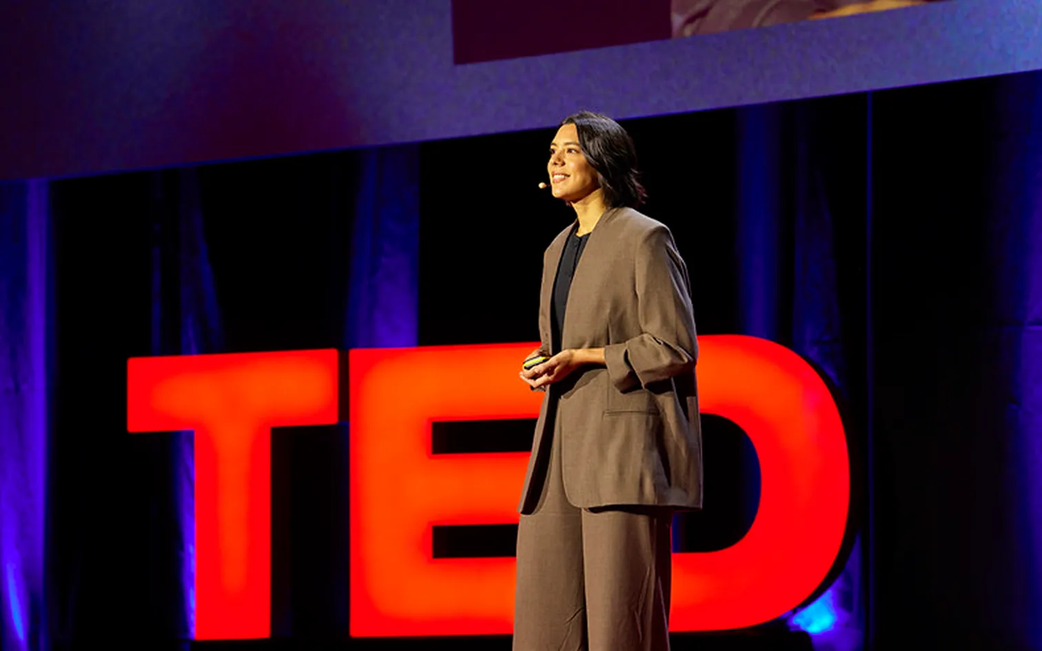 Woman in a brown suit speaking on stage with a large red TED logo behind her.