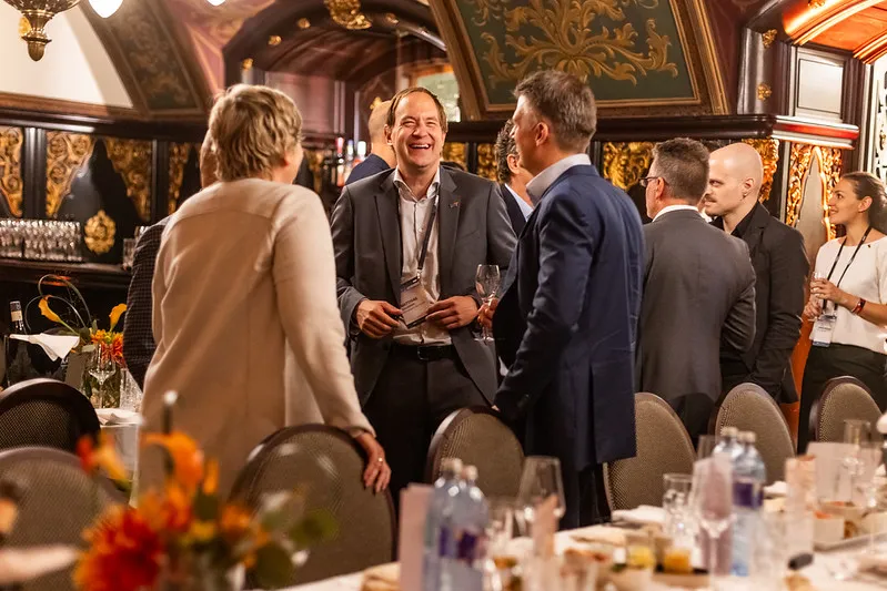 Group of professionally dressed people socializing and laughing in an ornate room with tables set for a formal event.