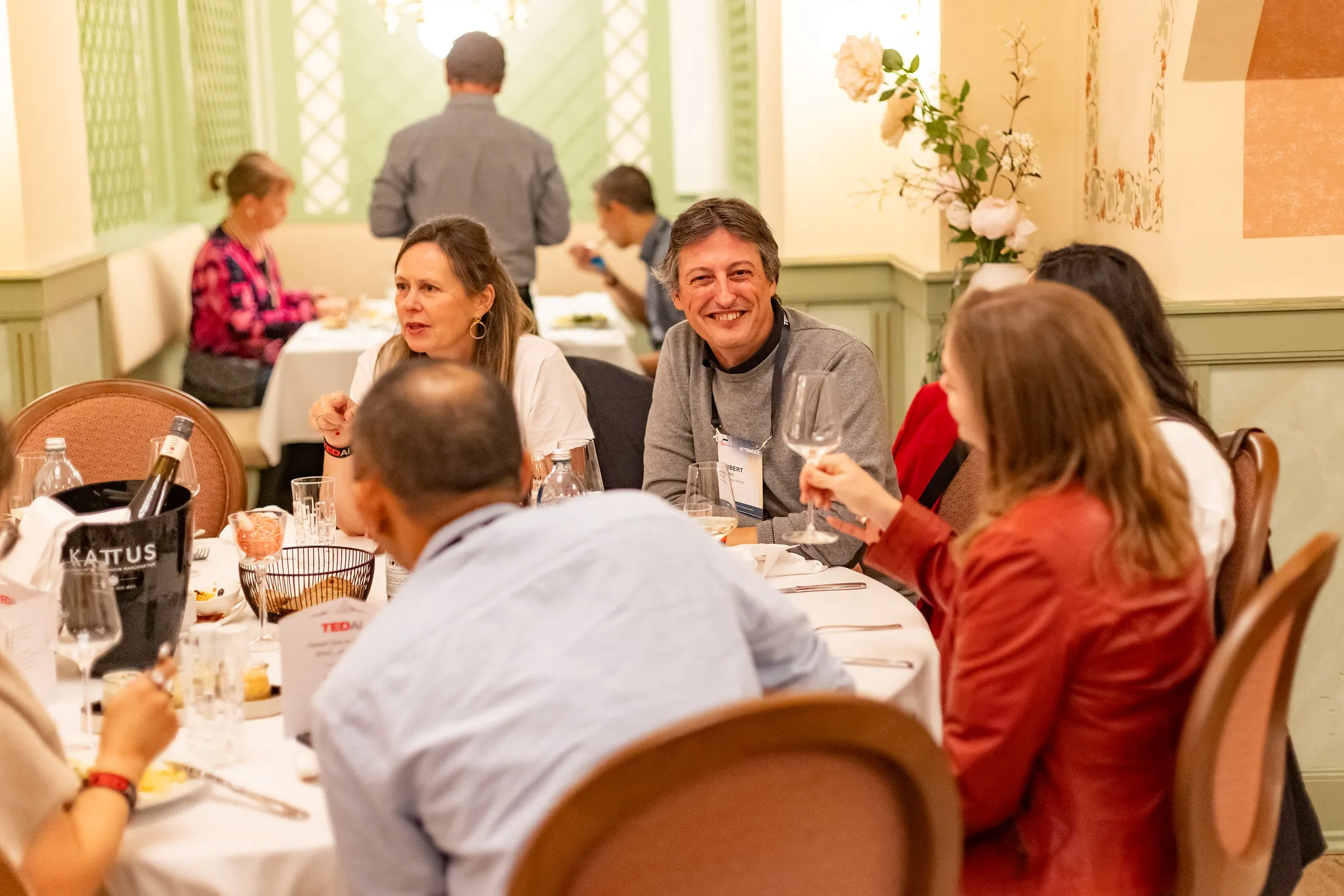 Group of people sitting around a round table having conversation and drinks in a warmly lit room.