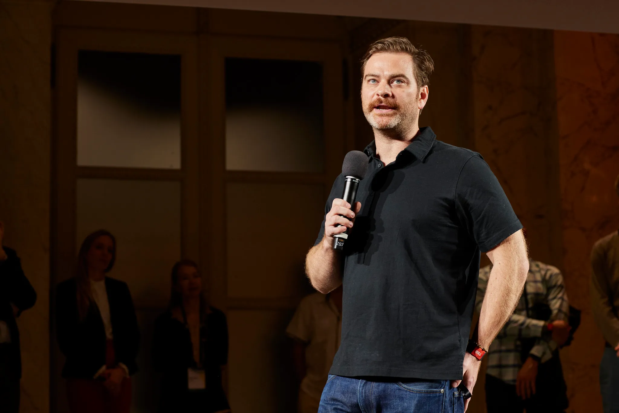 Man holding a microphone speaking on stage with people blurred in the background.