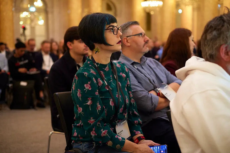Woman with short black hair and glasses attentively listening at a conference, holding a phone in her hands.