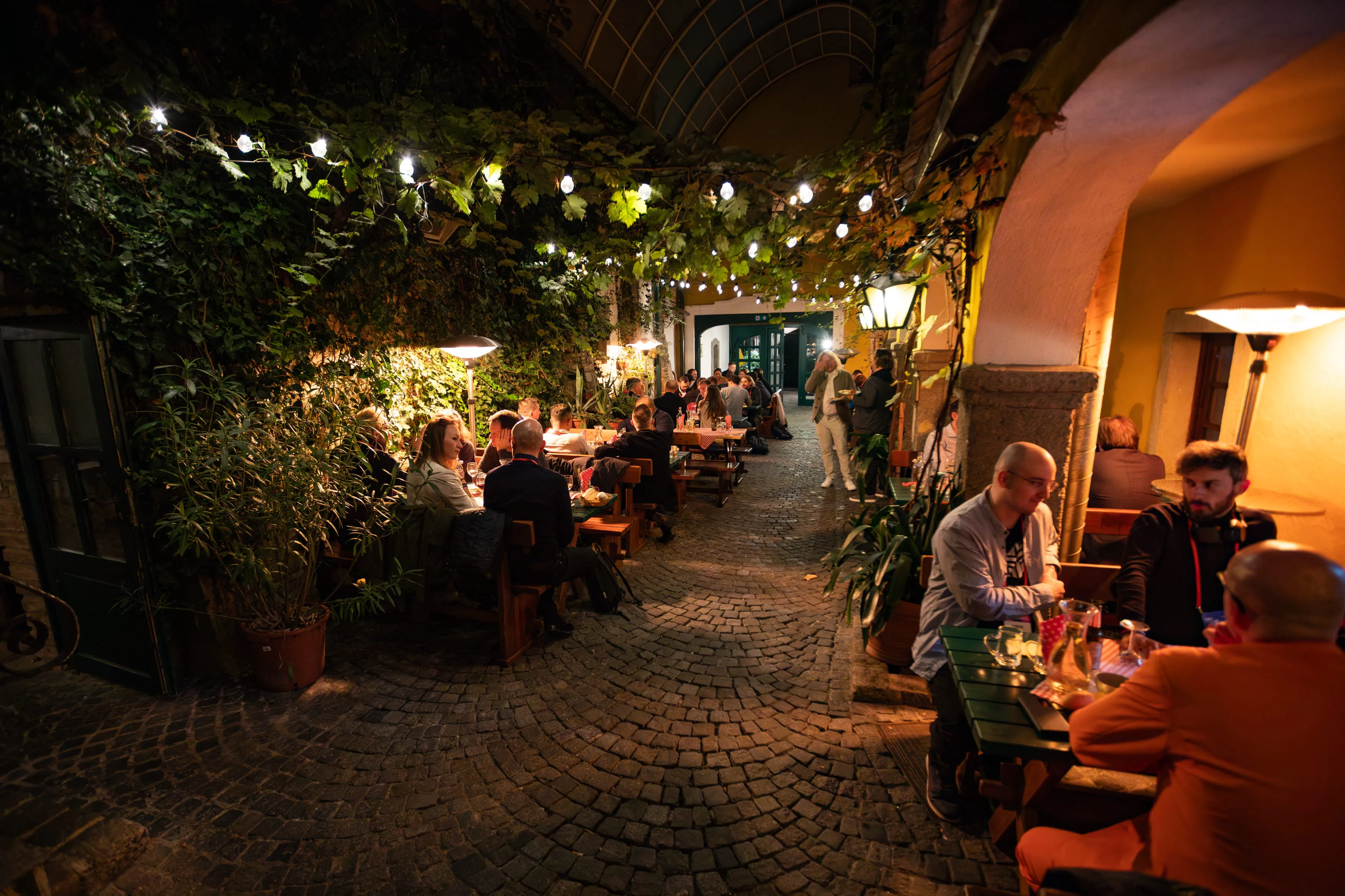 People dining and socializing at tables along a cobblestone alley decorated with string lights and greenery at night.