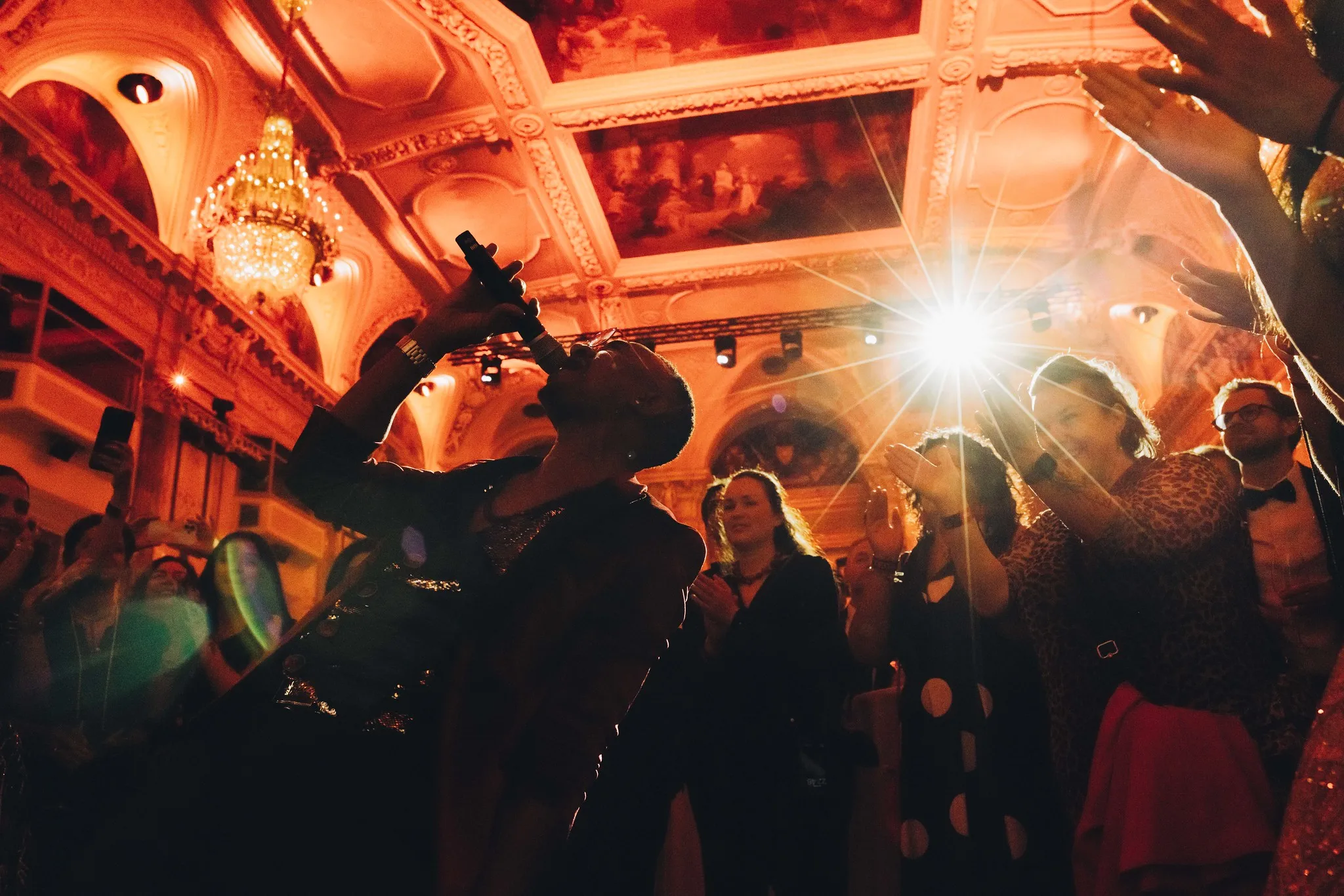 Singer passionately performing with a microphone in an ornate hall while an audience claps and cheers.