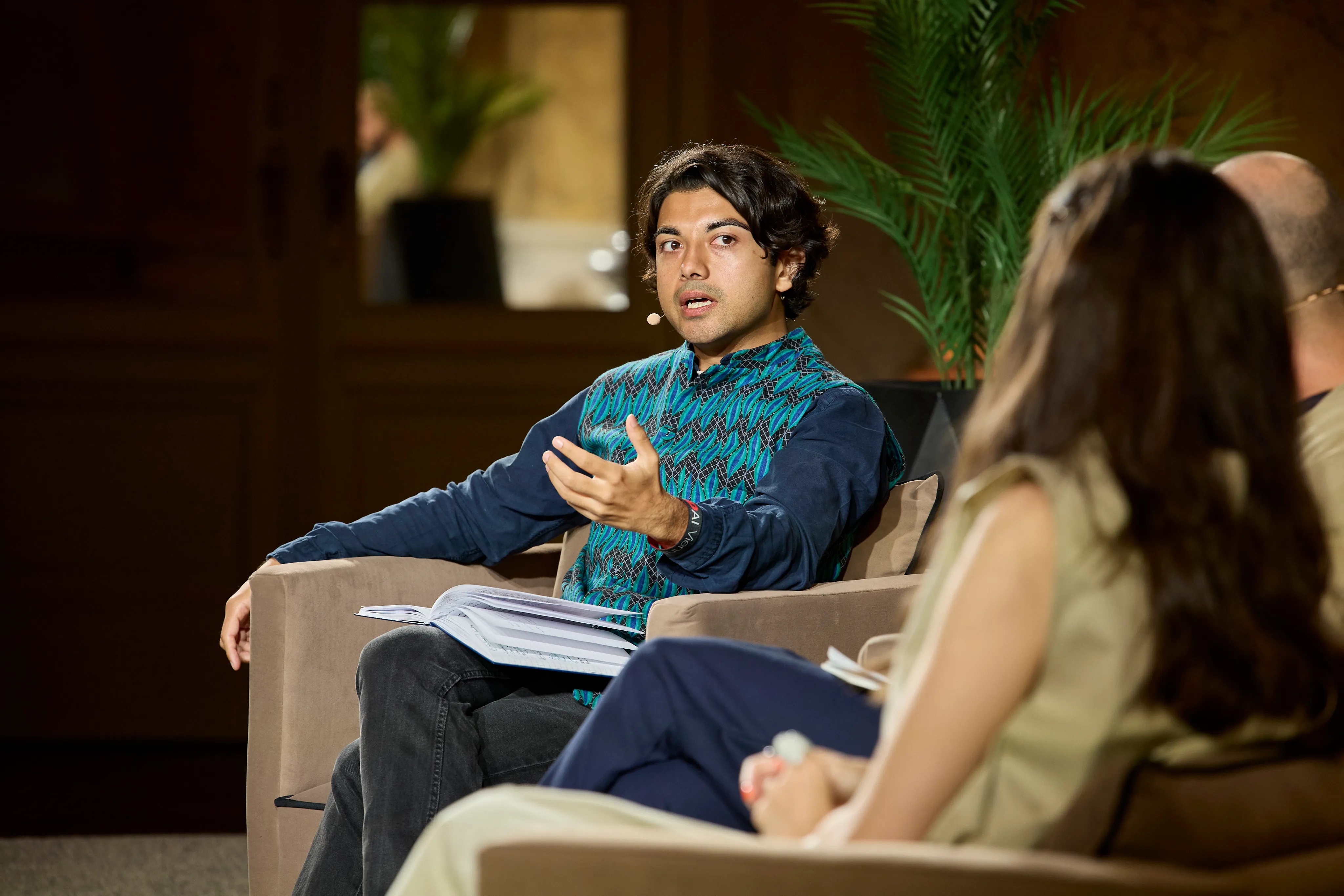 Man in a patterned blue vest speaking while seated with an open book on his lap during a discussion.
