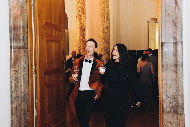 Man in a brown tuxedo and woman in a black dress holding drinks, walking through a doorway in a marble-paneled hall during an event.