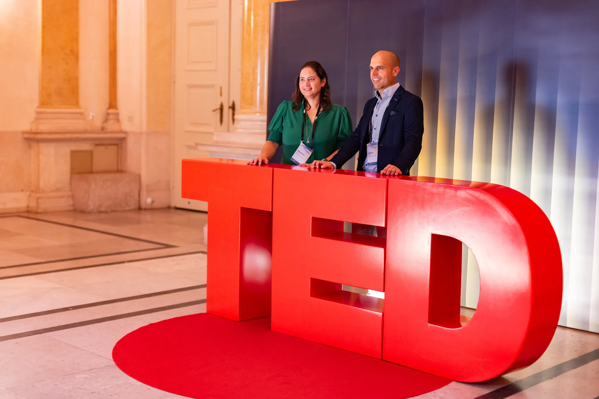 Two people standing behind large red TED letters on a red circular carpet in an elegant hallway.