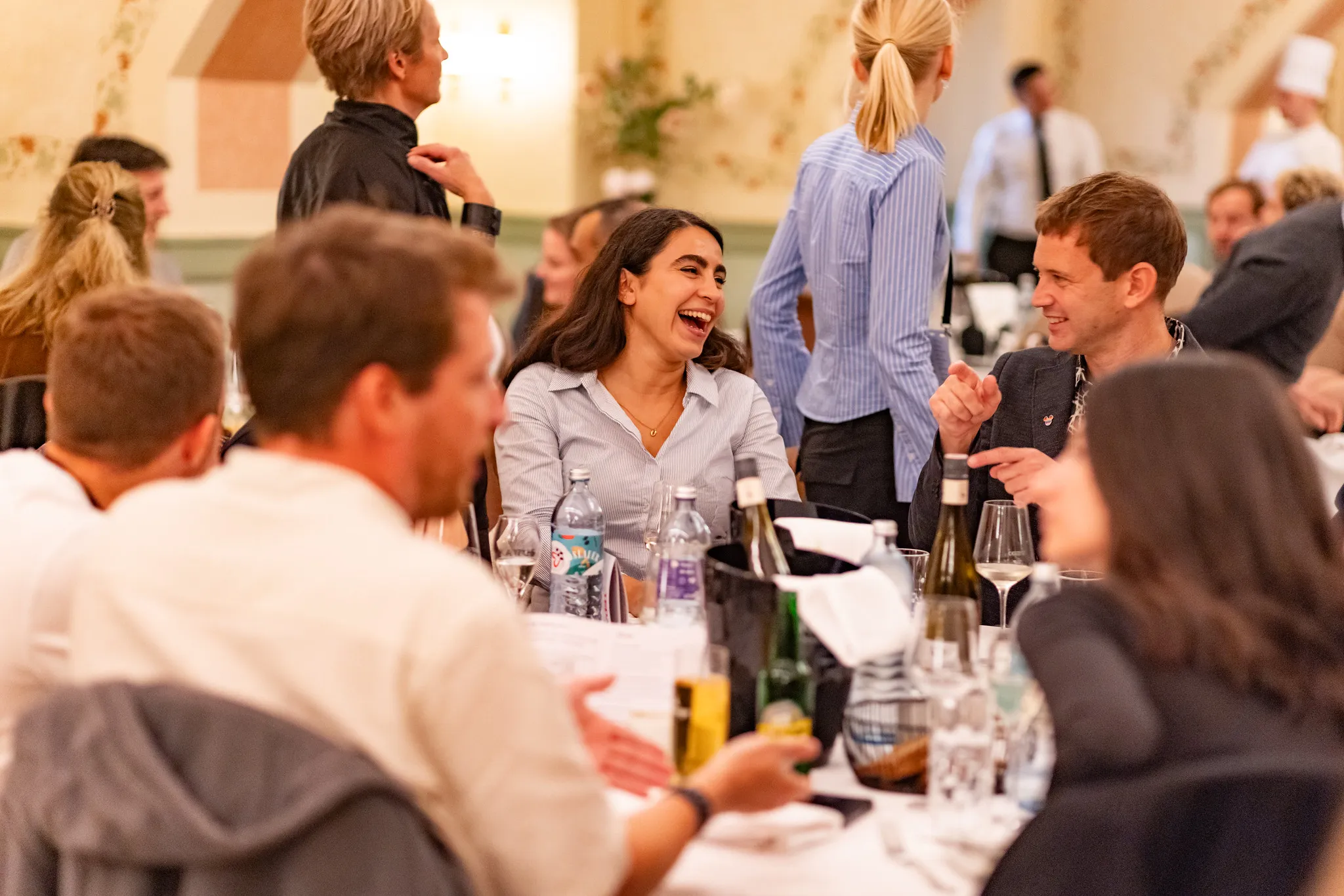 Group of people enjoying conversation and laughter around a dining table with drinks and wine bottles.