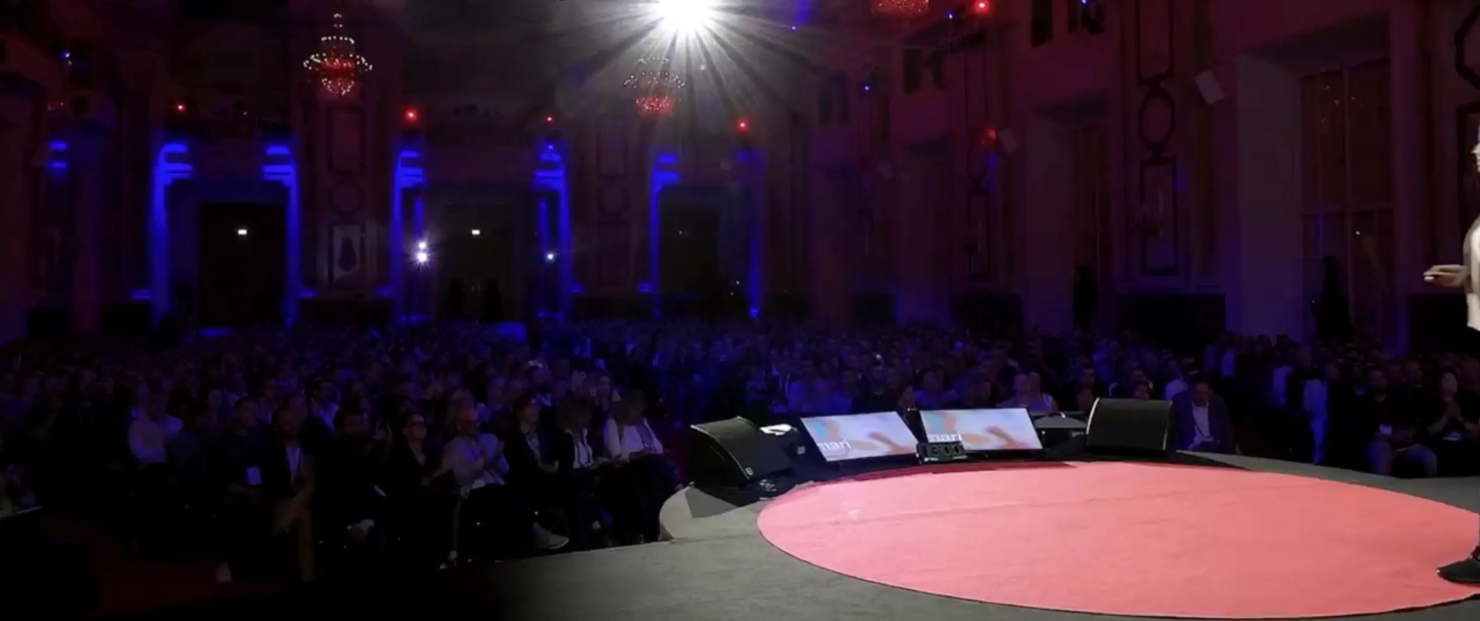 Large audience seated in a dimly lit auditorium facing a stage with a red circular carpet and a speaker partially visible on the right.