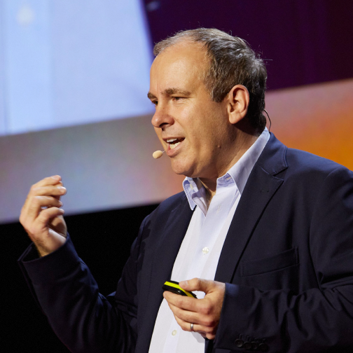 Man wearing a gray suit and white shirt speaking on stage with a headset microphone and gesturing with his hand.