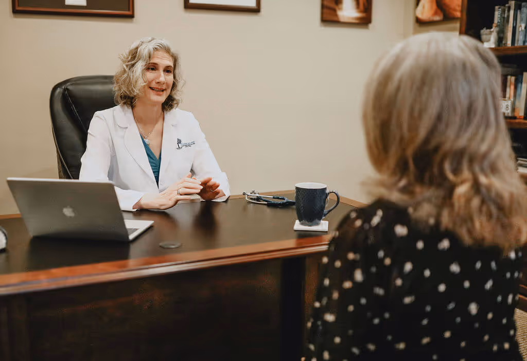 Dr. Libert sits smiling at her desk in a white lab coat, talking with a woman patient who's back is to the camera