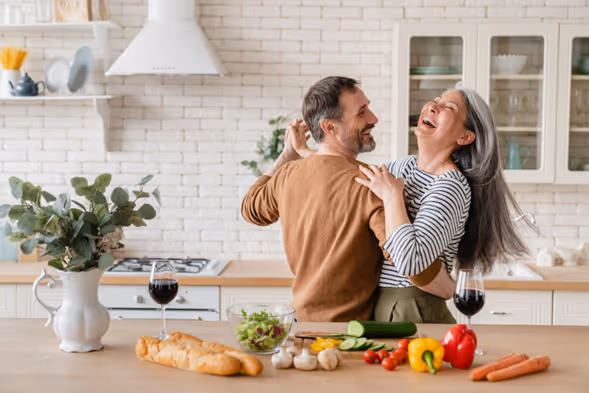 a grey haired woman laughs as she dances with her greay haired husband in the kitchen.  There is a variety of fresh veg on the counter in front of them as fun interrupted meal prep. 