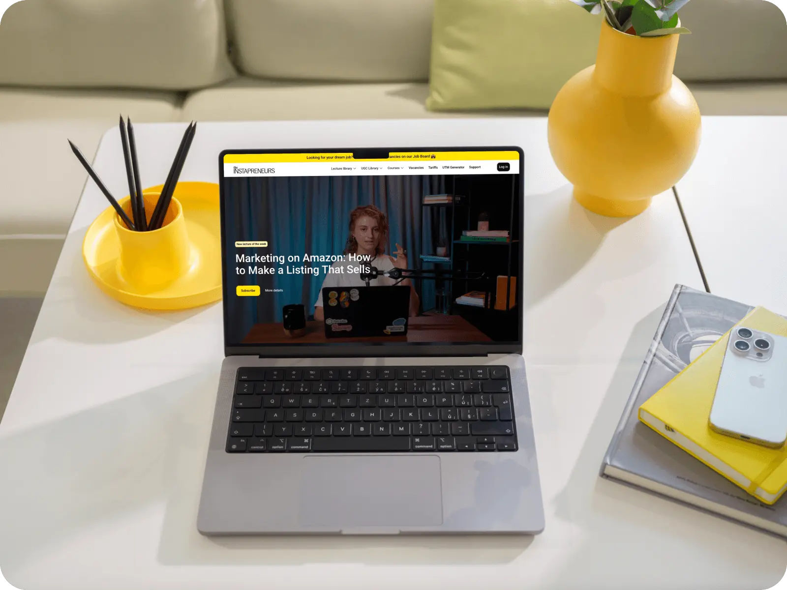 Open laptop on white table displaying a website about marketing on Amazon, with a yellow vase, pencil holder, and stacked books with an iPhone nearby.