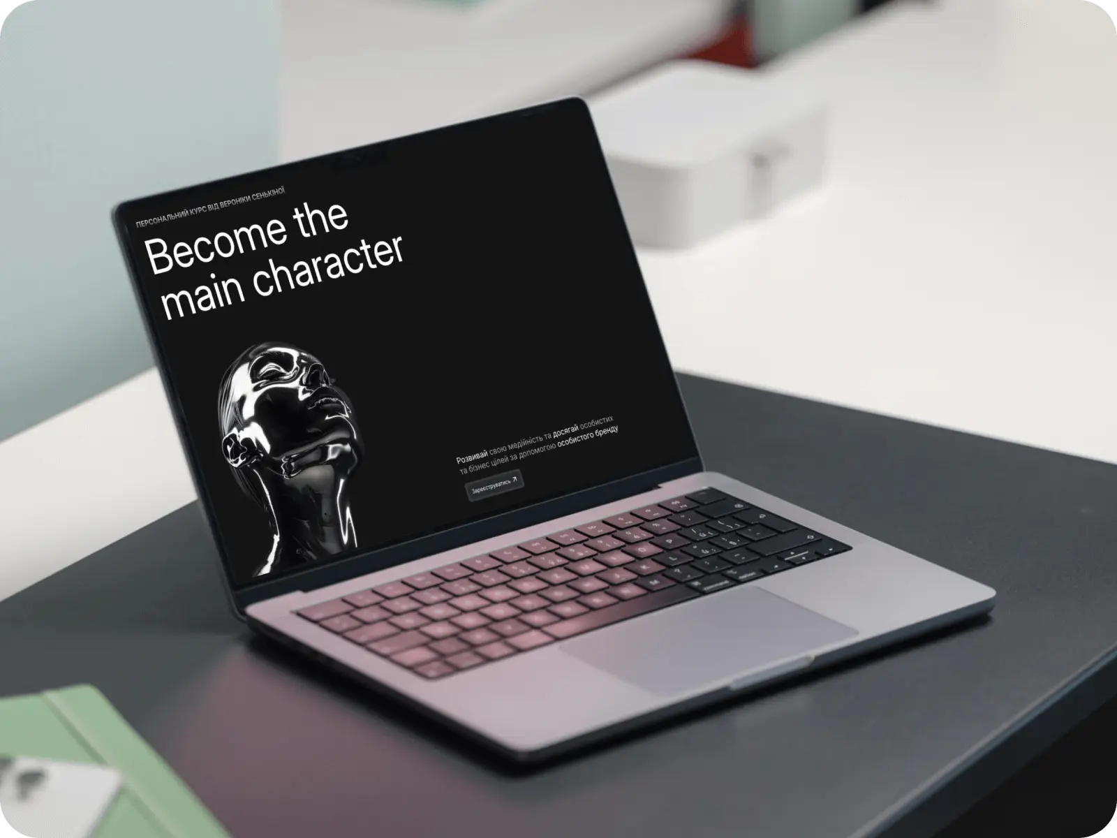 Laptop on a dark desk displaying a black screen with white text 'Become the main character' and a shiny black sculpture of a human head.