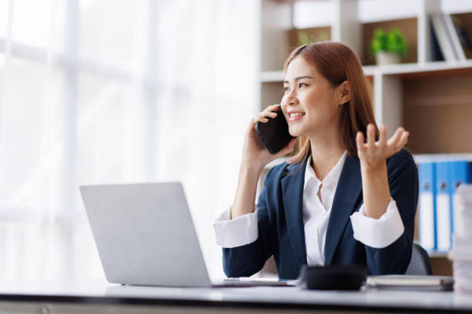 Smiling businesswoman in a navy blazer talking on a smartphone at her desk with a laptop and books nearby.